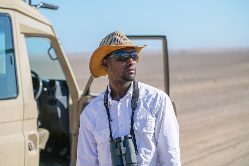 African man standing by safari vehicle wearing hat and white shirt and holding binoculars scans landscape looking for wildlife